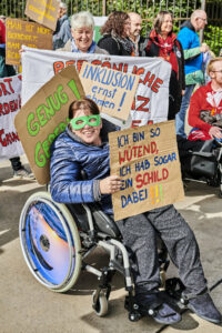 Eine Frau im Rollstuhl ist bei einer Demo und hält ein buntes Schild auf dem steht: "Ich bin so wütend, ich hab sogar ein Schild dabei!"